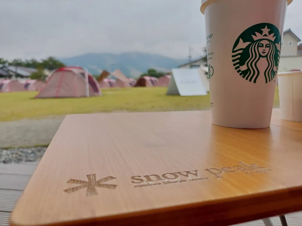 スノーピークの竹製ローテーブルとスタバのカップ、奥に広がる野外テントと山並み